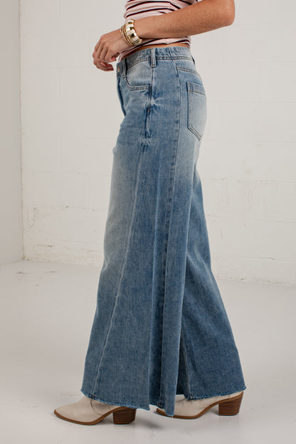 A person wearing high-waisted, wide-leg blue denim jeans with a faded wash, paired with a striped top and beige heeled boots, shown from the waist down in a bright studio setting.