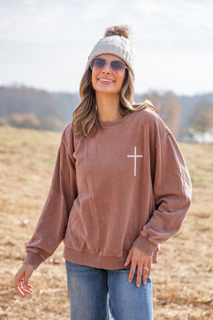 Woman wearing a brown sweatshirt with a white cross on it, standing in an open field.