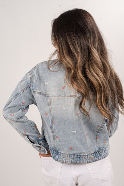 Person wearing a light blue denim jacket with floral embroidery on a white background