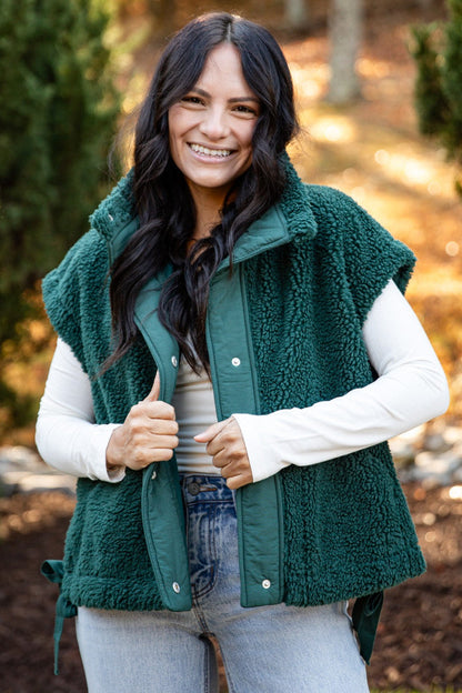 A woman wearing a dark green sherpa fleece vest over a white long-sleeve shirt and light blue jeans, standing outdoors in a park-like setting with trees and autumn foliage in the background.