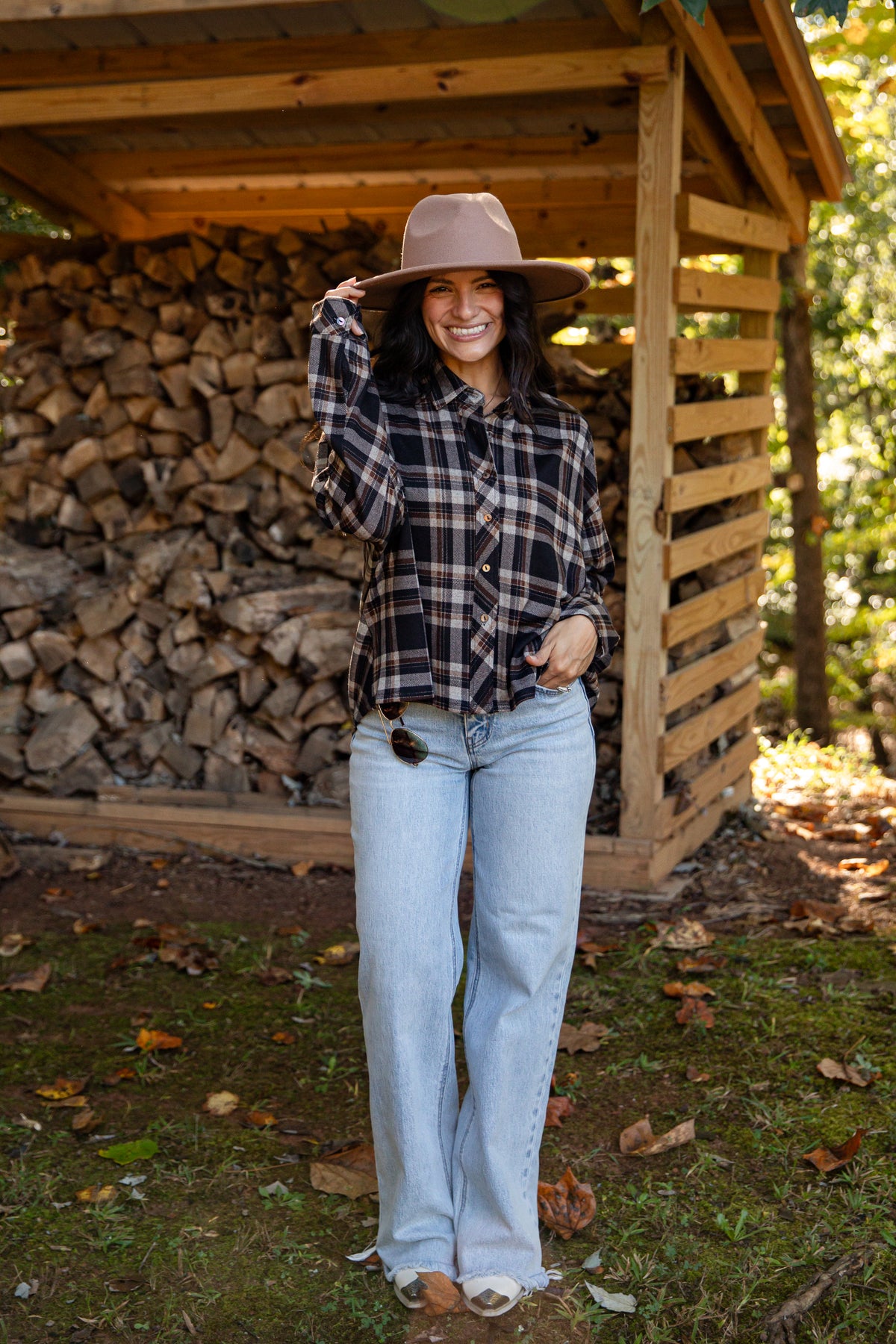 A woman stands outdoors in front of a wooden firewood shed, wearing a brown wide-brim hat, a black and white plaid button-up shirt, light-wash blue jeans, and white sneakers.