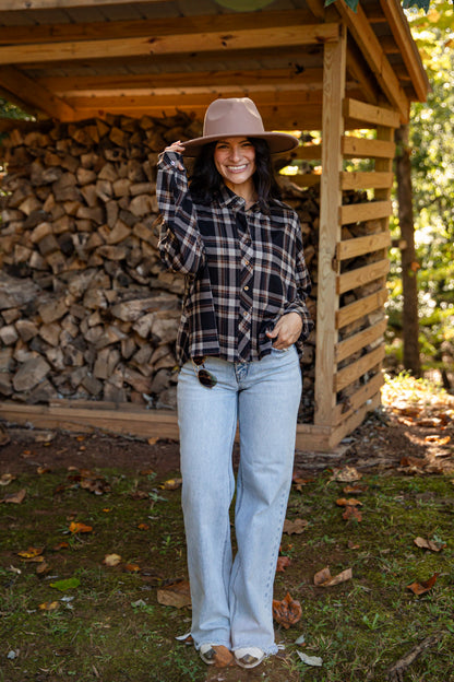 A woman stands outdoors in front of a wooden firewood shed, wearing a brown wide-brim hat, a black and white plaid button-up shirt, light-wash blue jeans, and white sneakers.