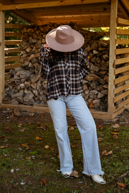 A person standing outdoors in front of a wooden firewood shelter, wearing a wide-brimmed dusty pink hat, a black and white plaid flannel shirt, light-wash blue jeans, and white shoes.
