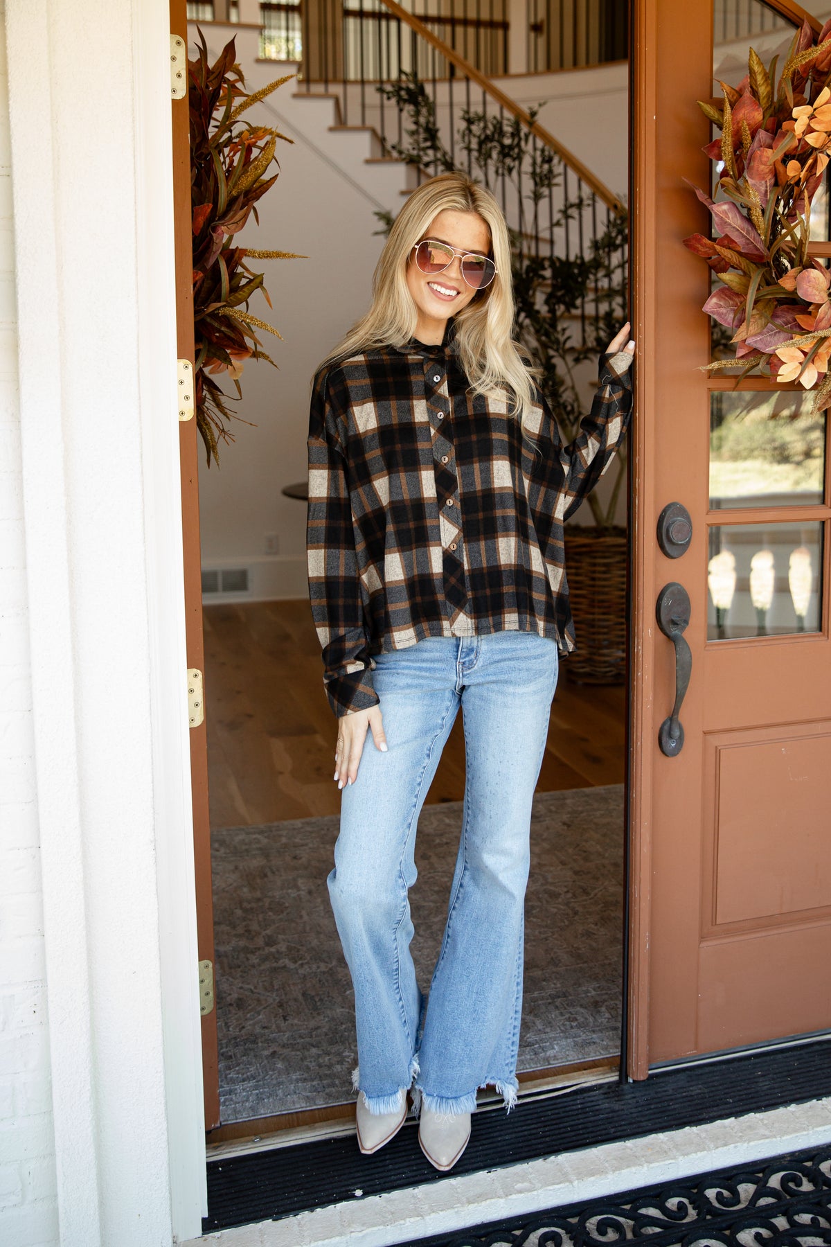 A woman stands in a doorway wearing a black and tan plaid button-up shirt, light blue flare jeans, and white pointed-toe heels. The setting is a home entryway with a brown door, wreaths, and a staircase in the background.