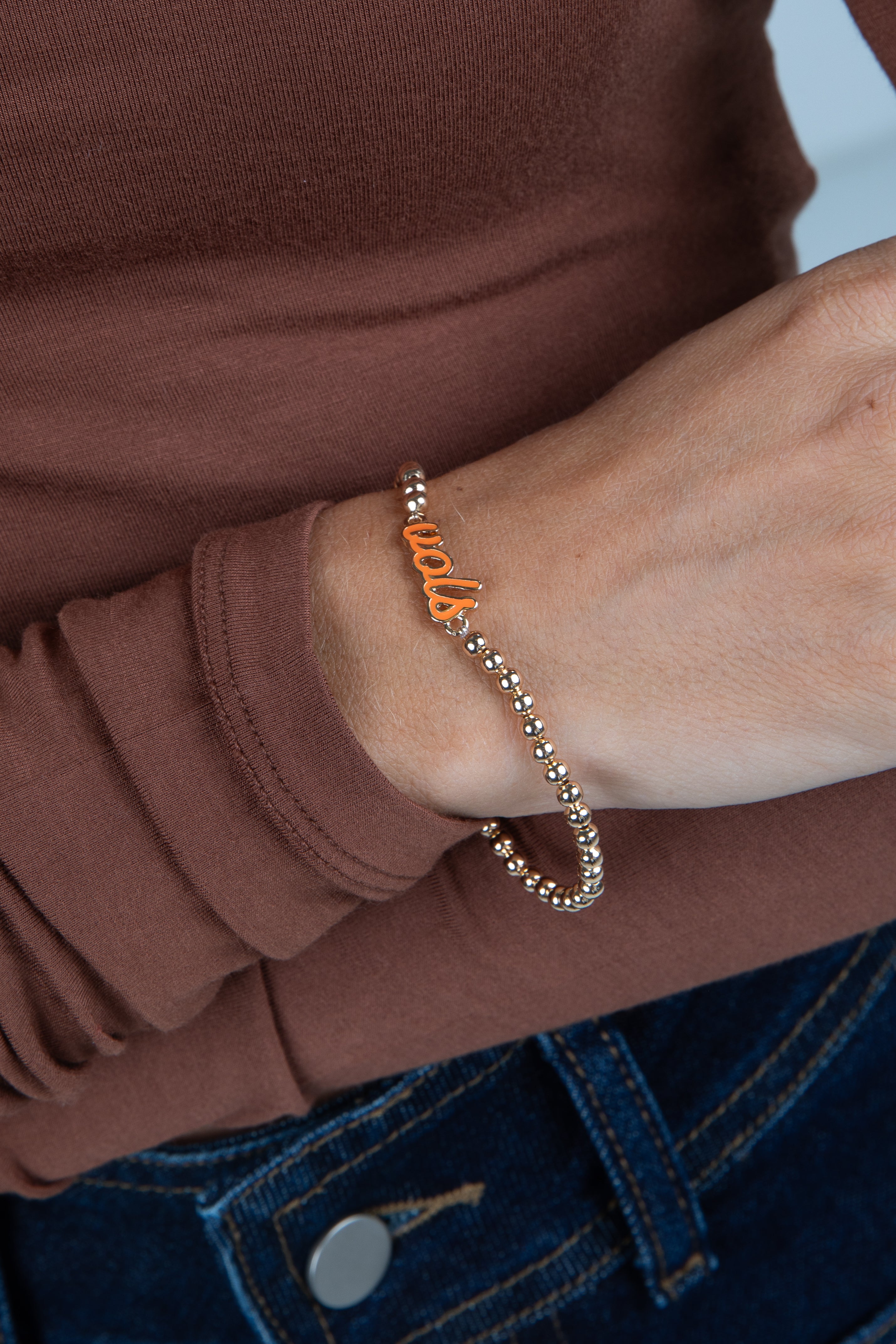 Close-up of a wrist wearing a bracelet with orange letters, against a brown shirt background.