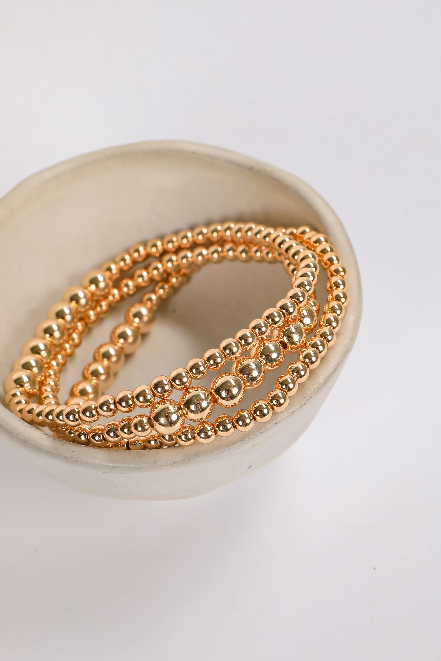 Three gold beaded bracelets on a white dish with a white background