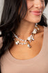 A close-up of a woman wearing a double-strand bohemian shell necklace with white pearls, seashells, and small gold-toned beads, paired with a beige scoop-neck top.