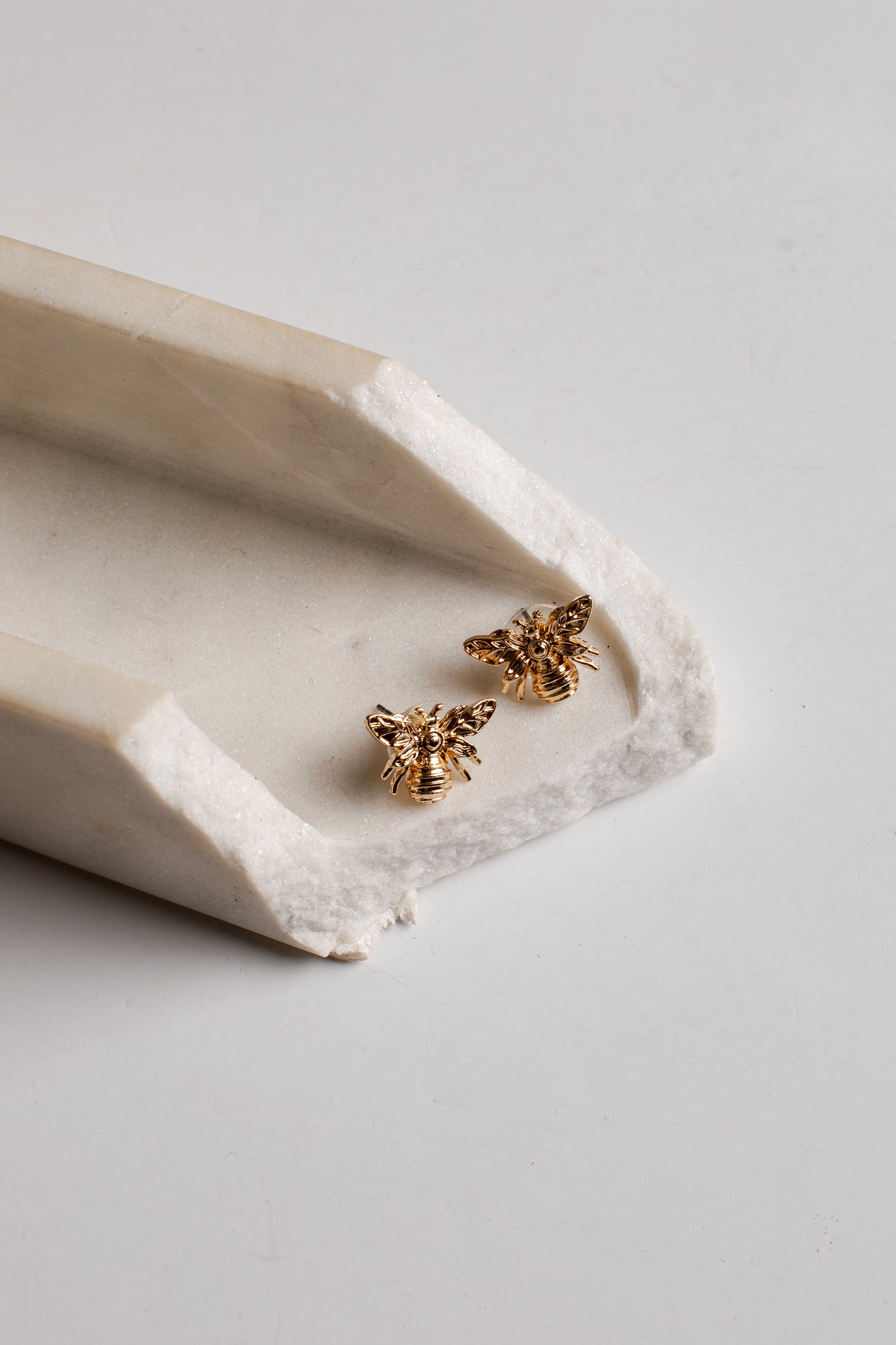 A pair of small gold bee stud earrings resting on a white ceramic tray against a light gray background.