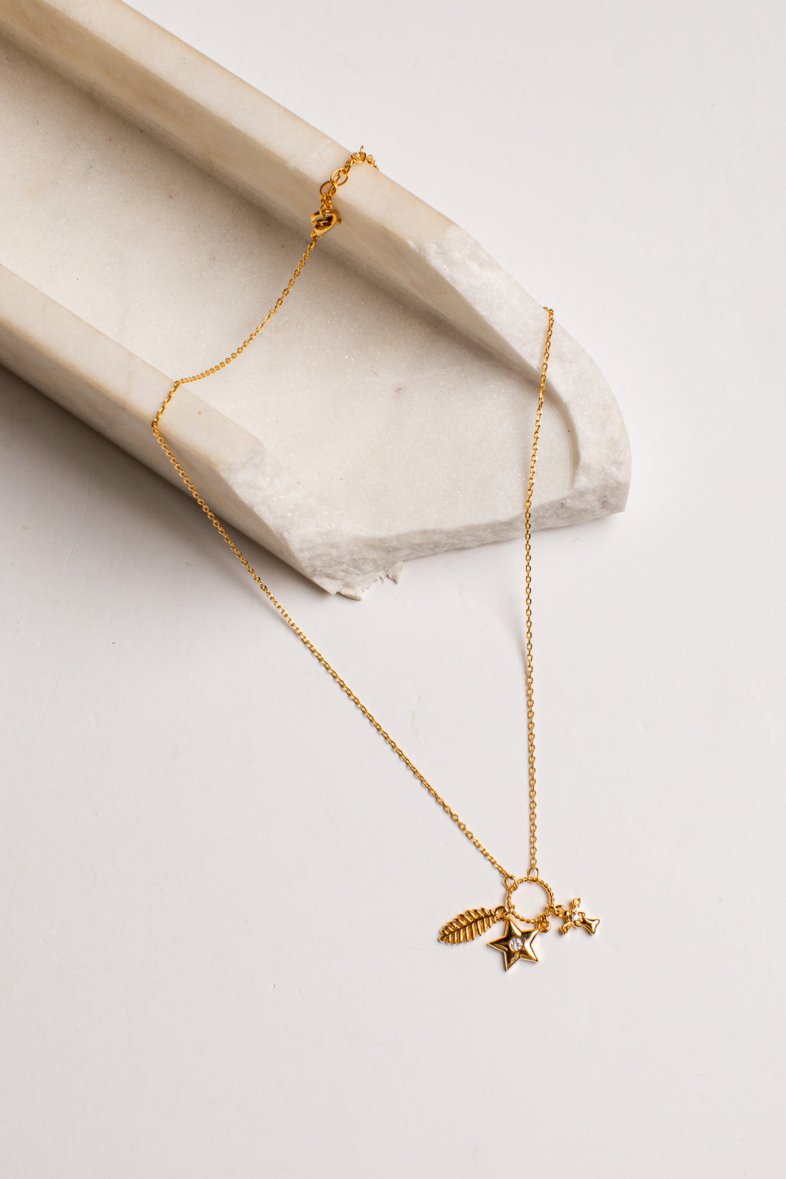 A delicate gold chain necklace with a small celestial pendant featuring a star, crescent moon, and feather, displayed on a light beige stone prop against a clean white background.