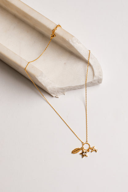 A delicate gold chain necklace with a small celestial pendant featuring a star, crescent moon, and feather, displayed on a light beige stone prop against a clean white background.