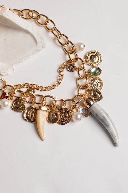 Gold-tone charm bracelet featuring a chunky chain, multiple coin charms, a cream-colored tooth, a white bone fragment, and small pearl beads on a light background.