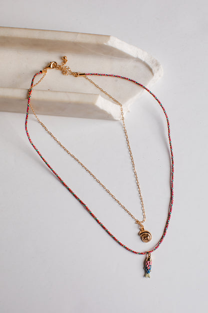 A delicate layered necklace set featuring a gold chain with a small gold spiral pendant and a longer red-and-gold beaded chain with a small multicolor fish charm, displayed on a light gray surface next to a white ceramic tray.