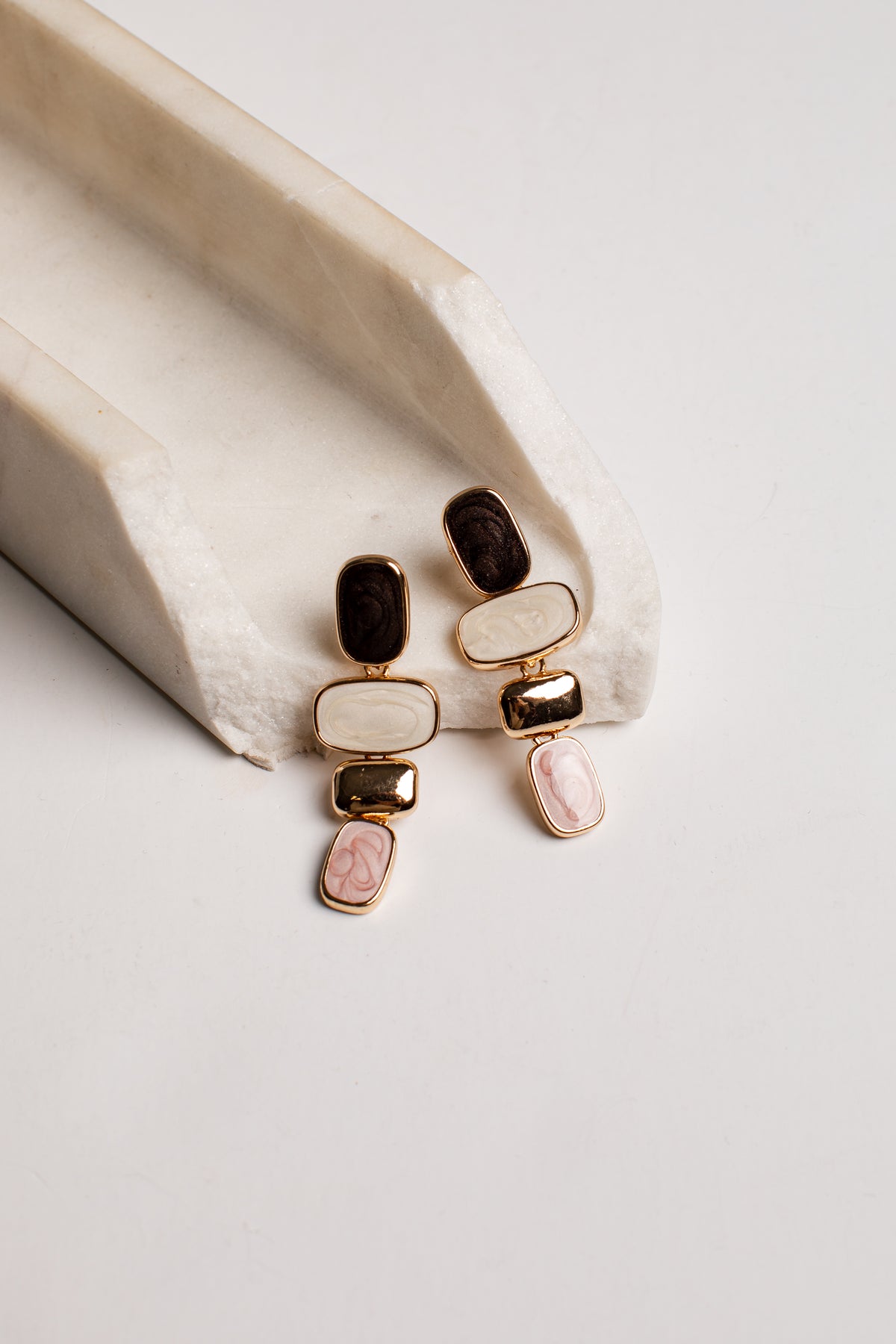 A pair of gold-toned statement earrings with stacked rectangular stones in cream, dark brown, and light pink, displayed on a cream-colored textured ceramic riser against a light gray background.