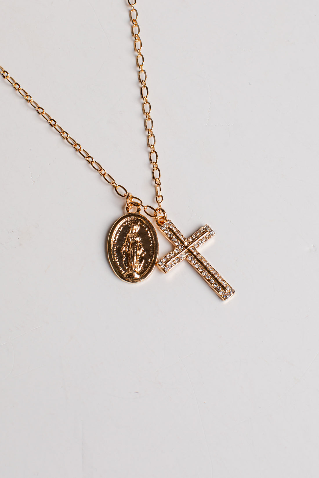 A rose gold chain necklace featuring two pendants: an oval Miraculous Medal pendant and a rectangular cross paved with small clear stones, displayed on a light gray background.
