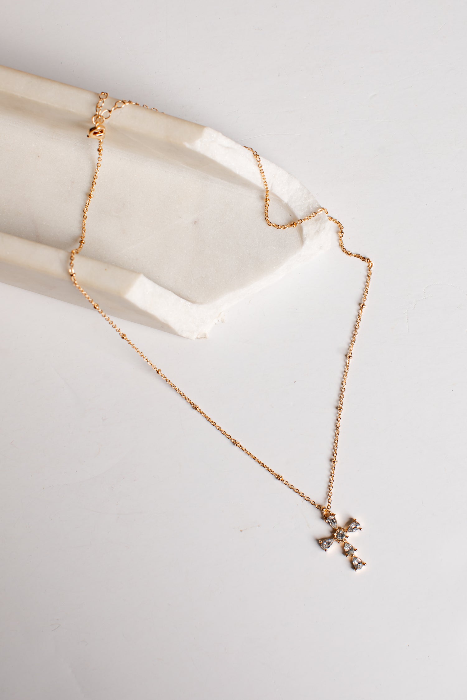 A delicate rose gold chain necklace with a small cross pendant, draped over a white stone display block on a light gray background.