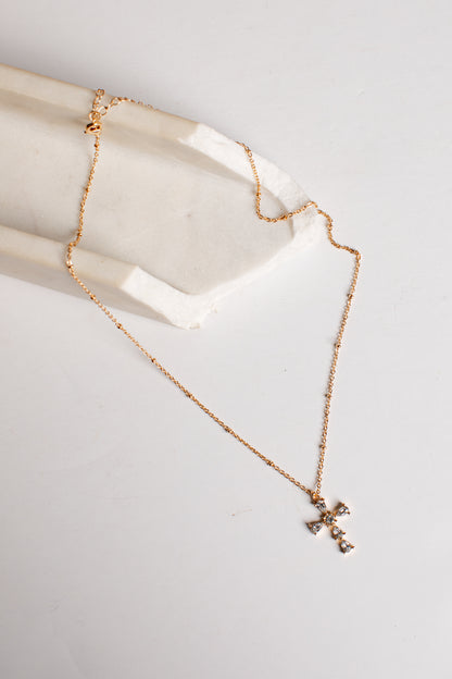 A delicate rose gold chain necklace with a small cross pendant, draped over a white stone display block on a light gray background.