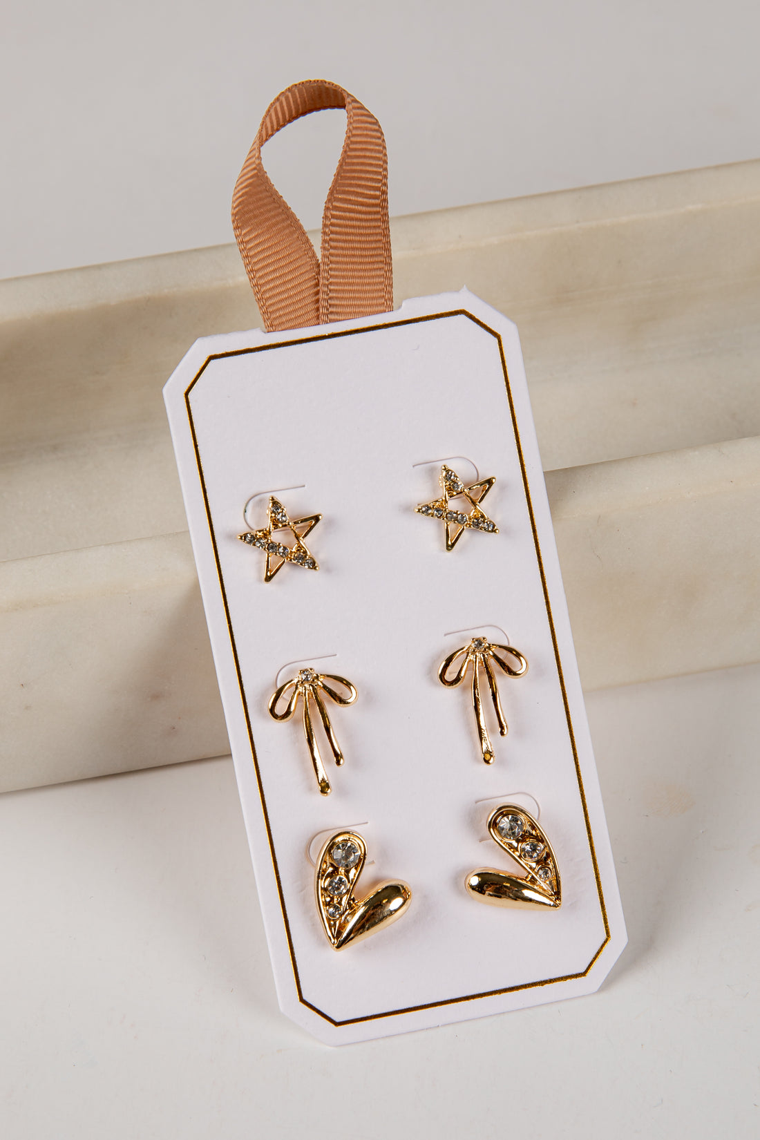 Set of gold earrings on a white card with a brown ribbon against a beige background