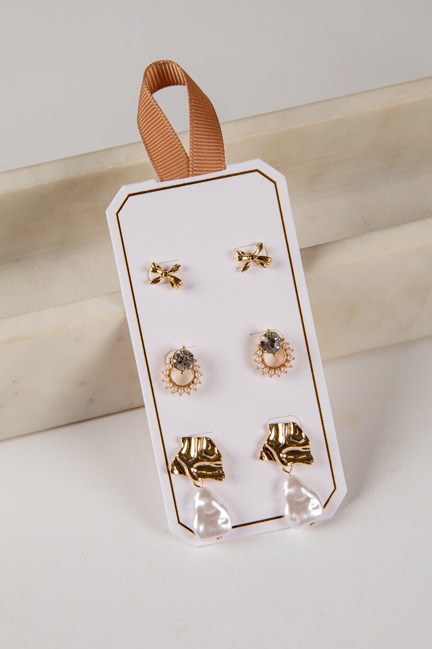 Set of gold earrings on a clear plastic card with a brown ribbon, against a light background.