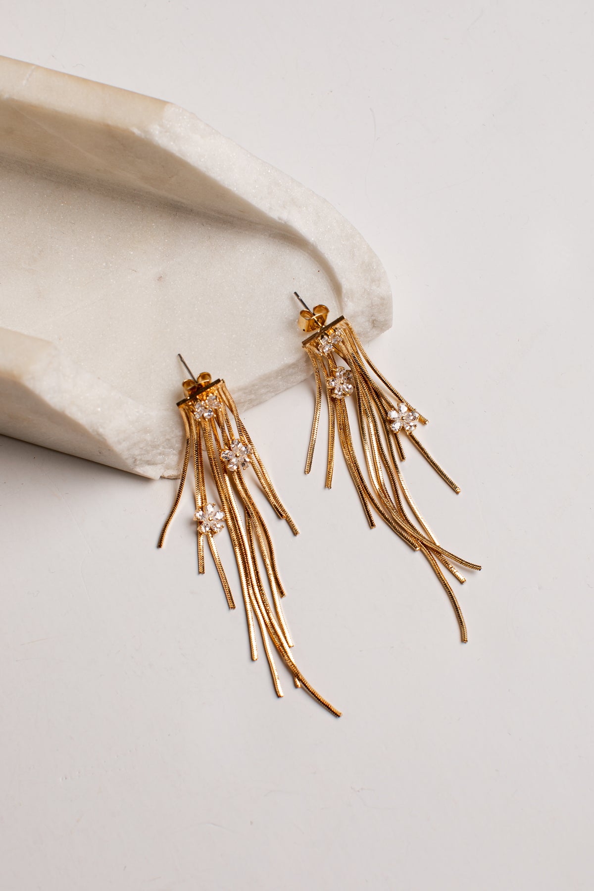 A pair of gold-toned dangling earrings with multiple thin fringe strands and small clear rhinestone clusters, displayed on a light neutral surface beside a curved cream stone prop.
