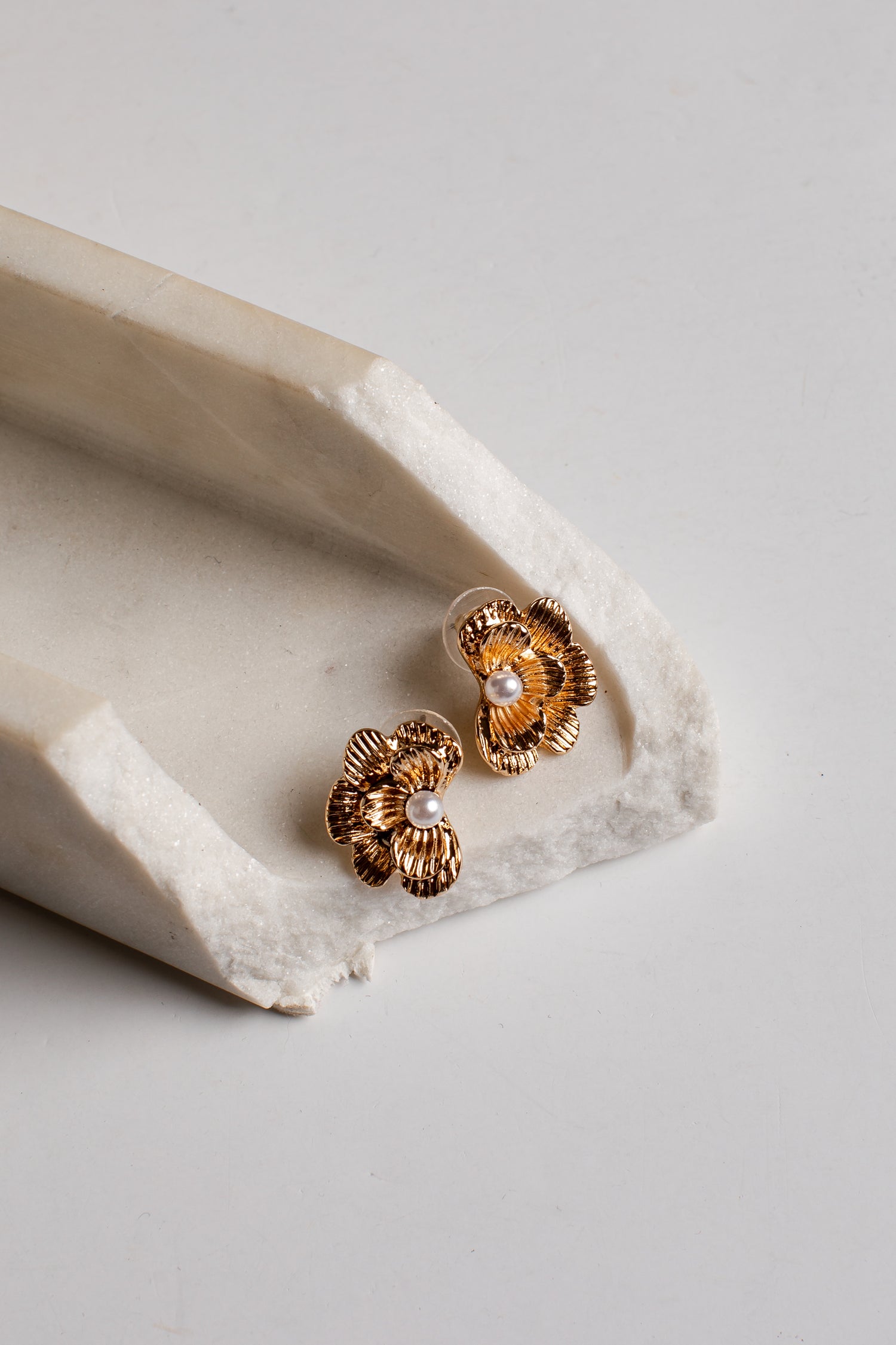 A pair of gold-toned flower-shaped stud earrings with small white pearl centers, resting on a textured off-white stone tray against a light gray background.