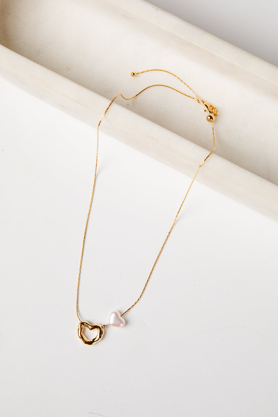 Gold necklace with a heart-shaped clasp and pearl pendant on a white surface