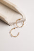 A pair of gold-toned open hoop earrings with small clear faceted beads, displayed on a light surface beside a cream-colored stone prop.
