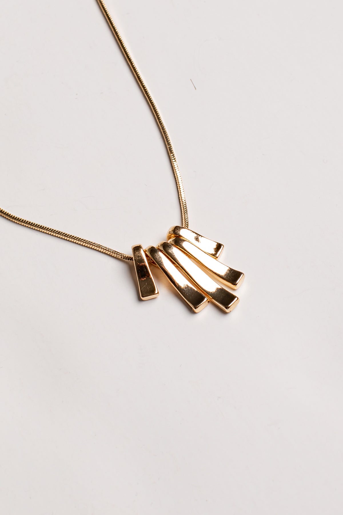 A gold necklace with a modern pendant featuring four overlapping rectangular bars, photographed on a clean, light off-white background.
