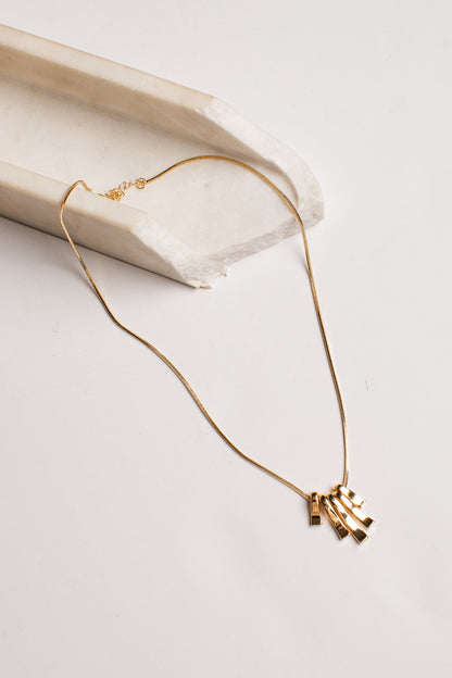 A delicate gold chain necklace with a small cluster of geometric bar pendants, styled on a light beige stone prop against a clean white background.