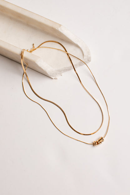 A delicate gold double-layer necklace with a small beaded accent on one layer, draped over a curved white stone prop on a light beige background.