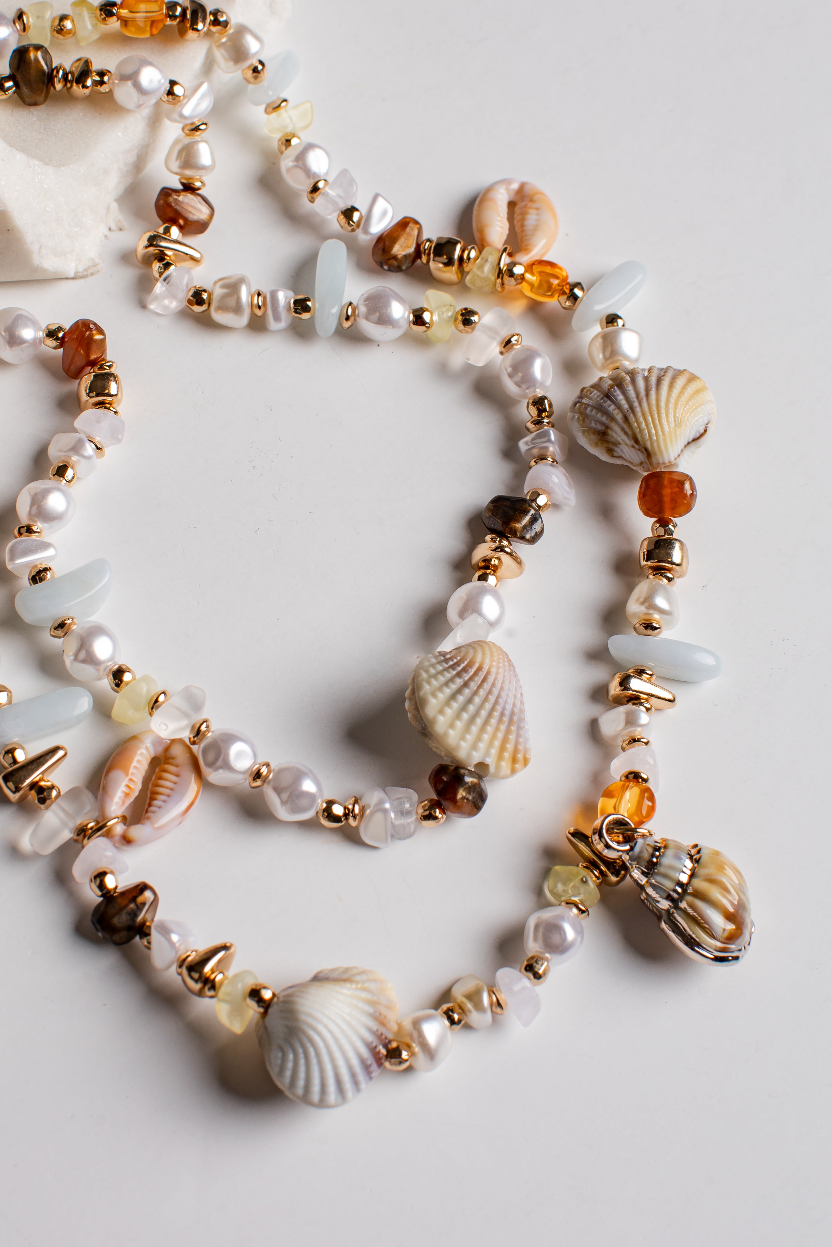 A multi-strand beaded necklace featuring white pearls, seashells, amber-colored beads, and gold-toned spacer beads, arranged on a light gray background.