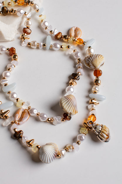 A multi-strand beaded necklace featuring white pearls, seashells, amber-colored beads, and gold-toned spacer beads, arranged on a light gray background.