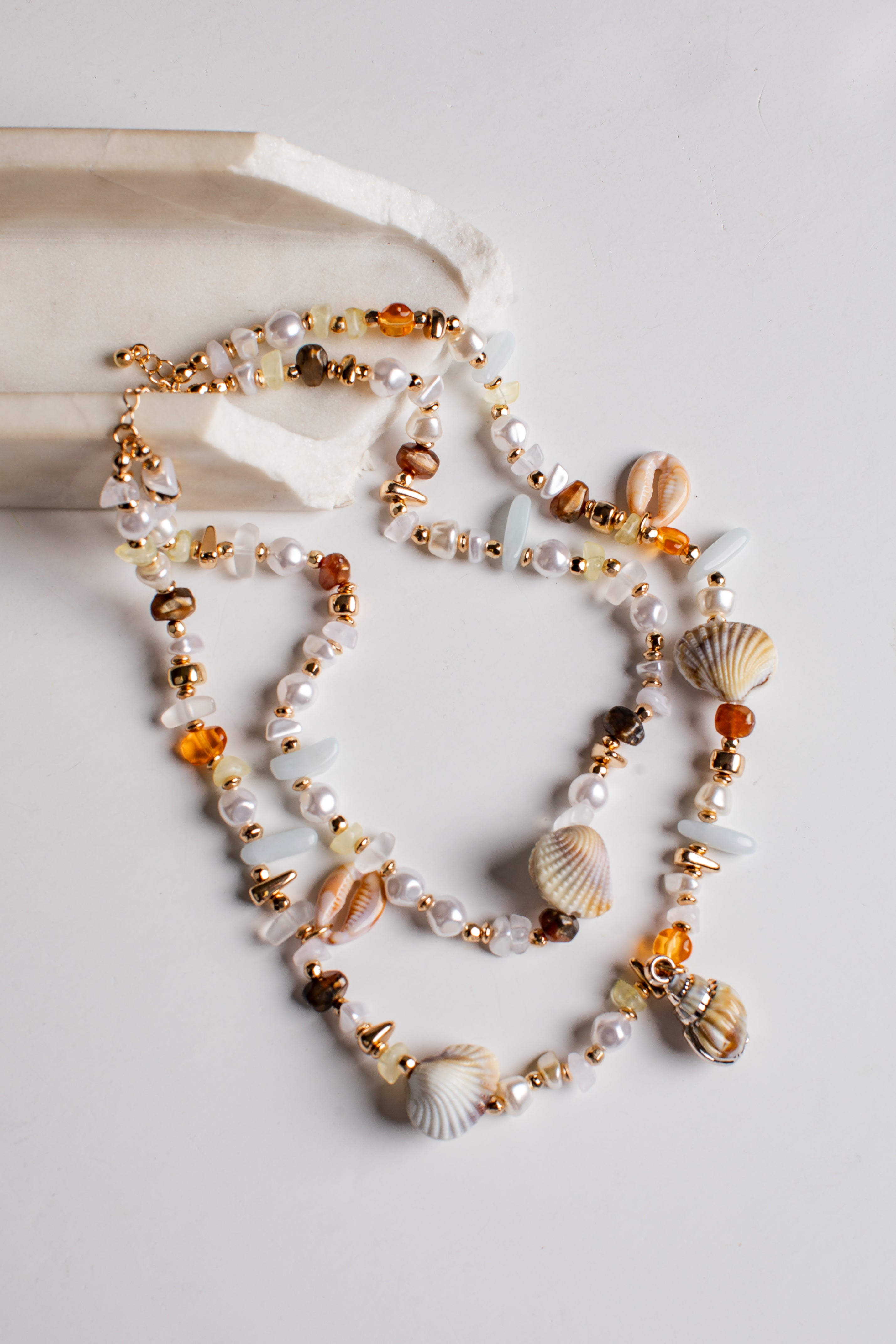 A multi-strand beaded necklace with seashells, white pearls, amber-colored beads, and gold-toned metal accents, styled on a light gray surface with a white stone prop.