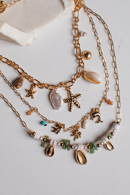 A layered gold-tone necklace set with seashell and starfish charms, displayed on a light gray surface with a textured white stone prop.