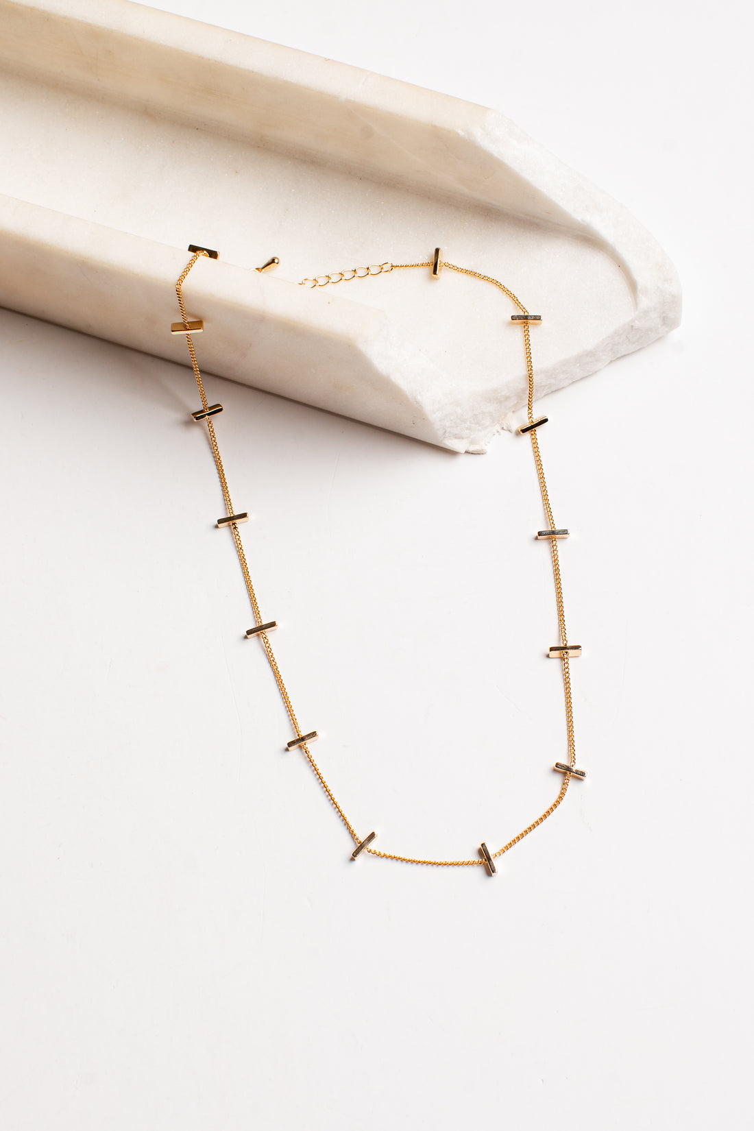 A delicate gold chain necklace with small, spaced cross-shaped links, displayed on a white surface next to a cream-colored stone block.