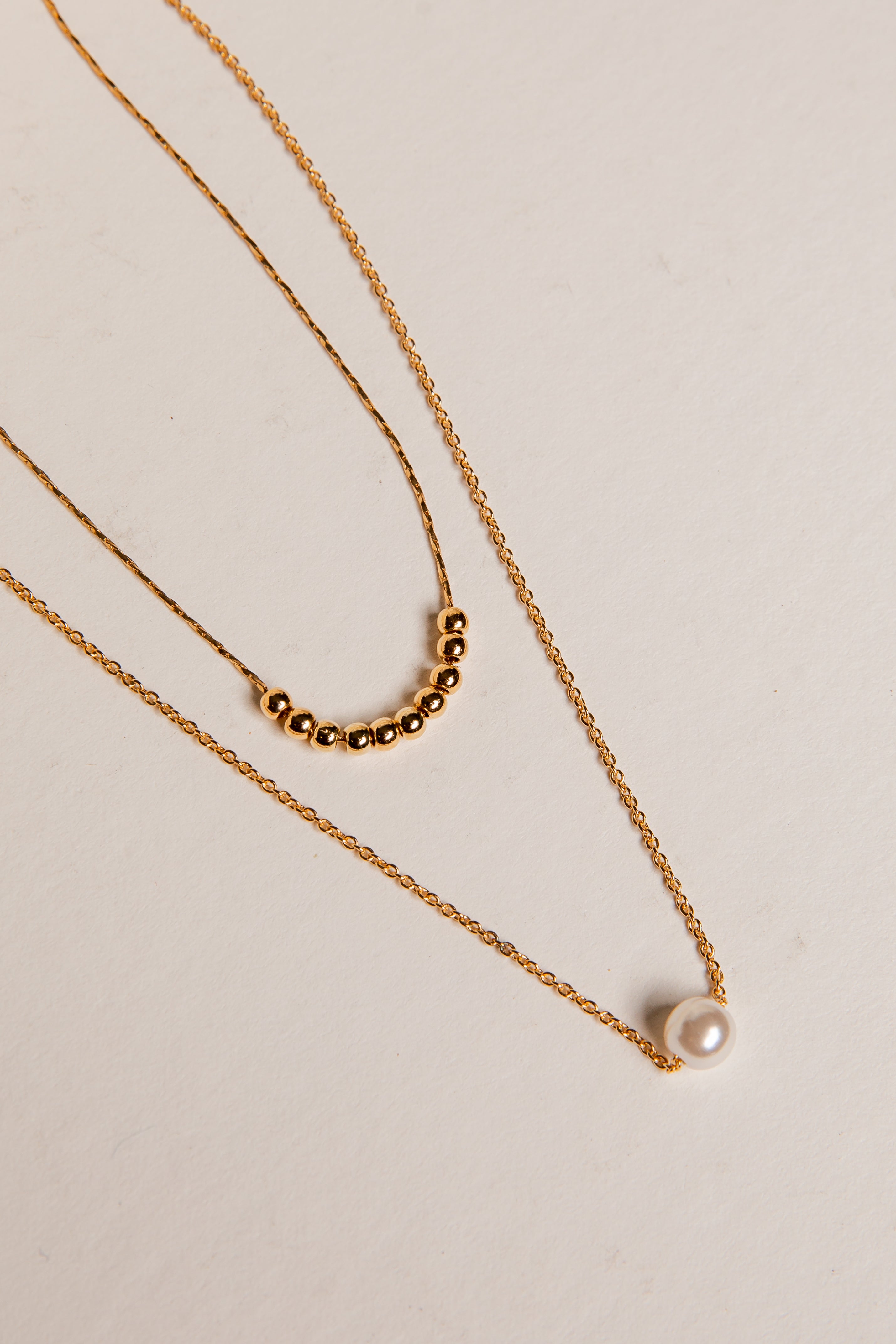 Two gold necklaces with beads and a pearl on a beige background