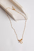 A delicate gold chain necklace with a small charm featuring a clear gemstone and a gold square pendant, displayed on a light beige stone tray against a white background.
