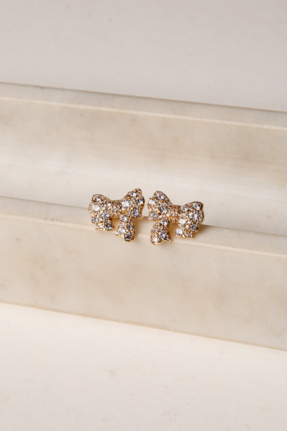 Gold bow-shaped earrings with rhinestones on a beige surface