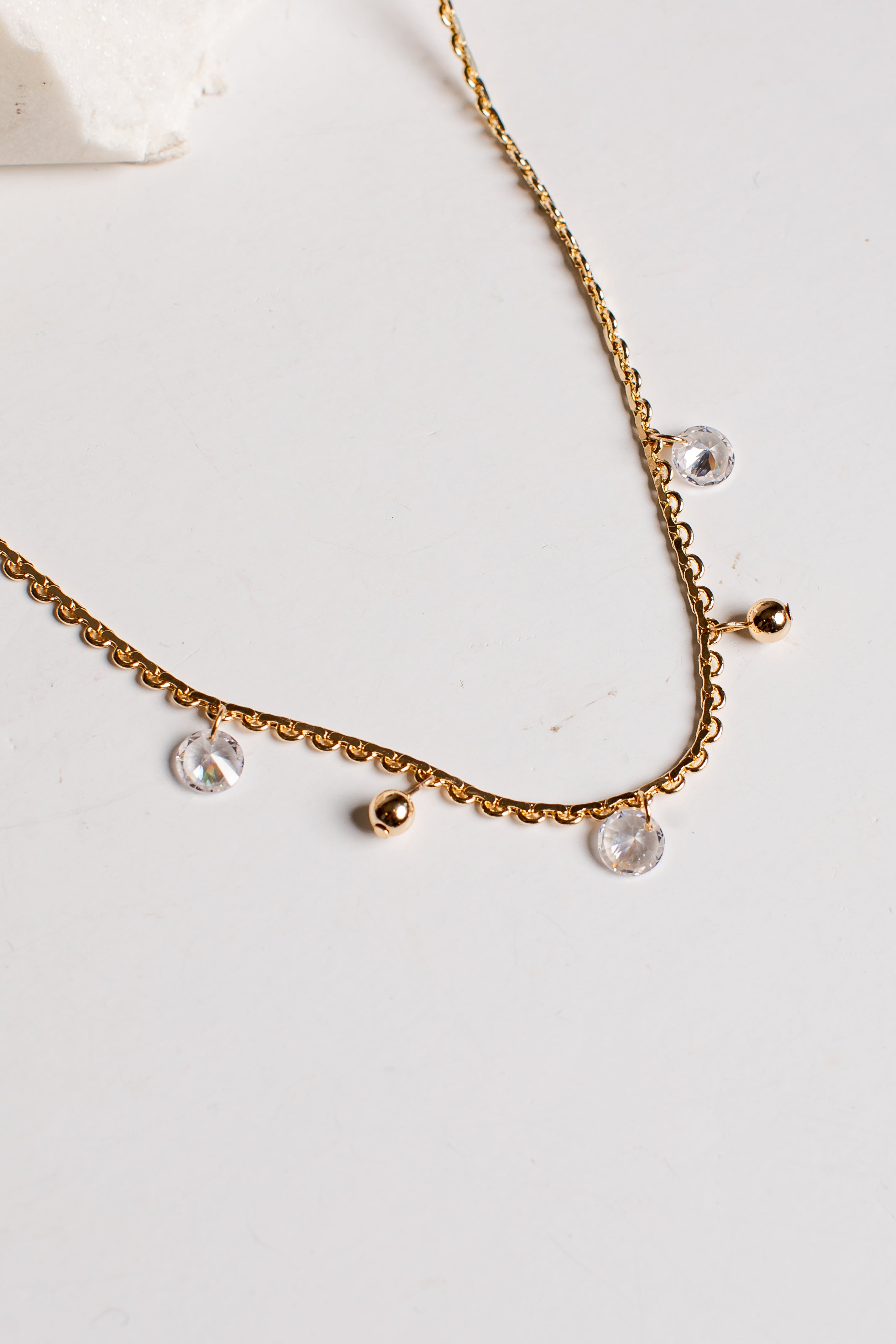 A delicate gold chain necklace with small gold beads and clear faceted crystal drops, displayed on a light off-white surface.