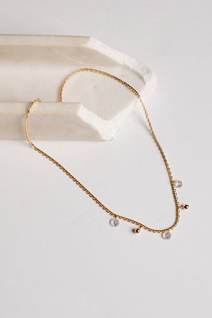 A delicate gold chain necklace with small clear crystal pendants and tiny gold beads, displayed on a white surface next to a cream-colored stone prop.
