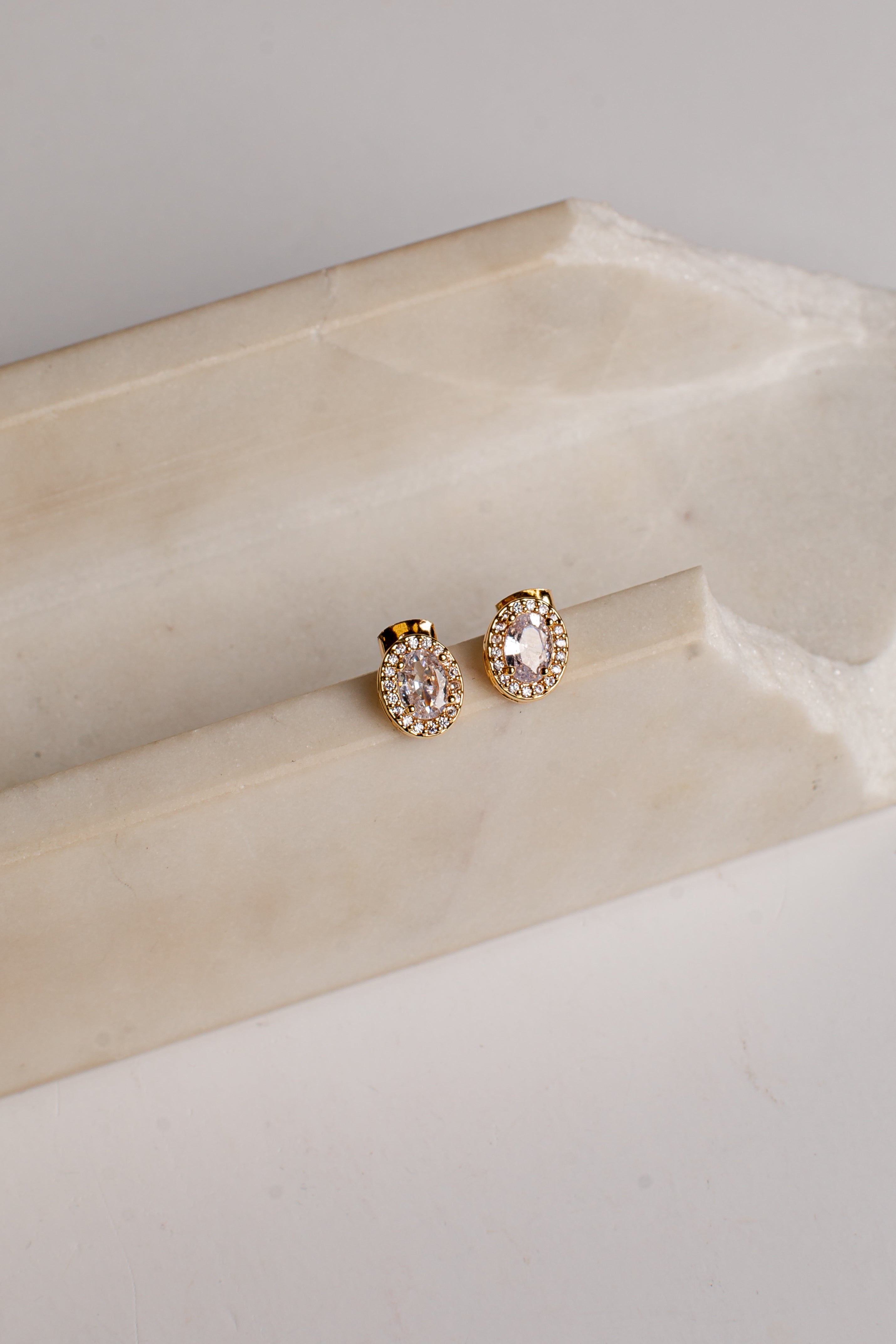 A pair of gold-toned stud earrings with oval clear gemstones and small surrounding clear stones, displayed on a cream-colored stone riser against a light neutral background.
