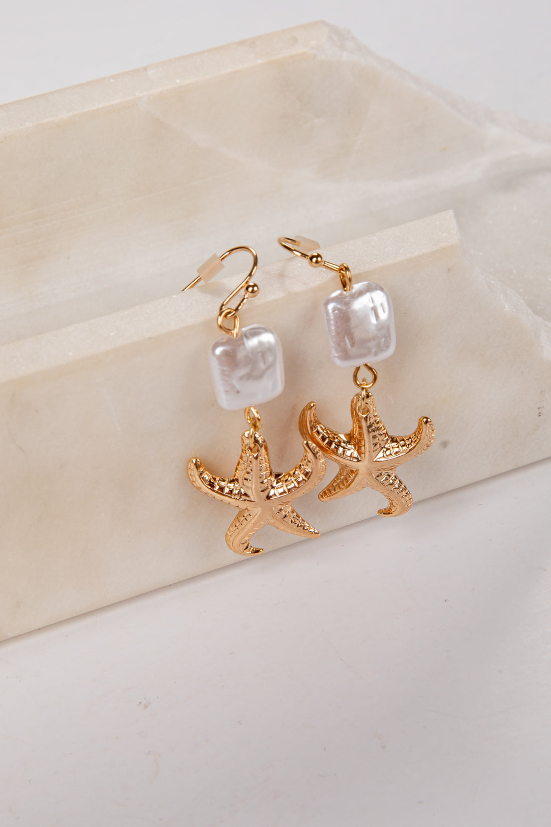Gold starfish earrings with pearl-like beads on a beige stone surface