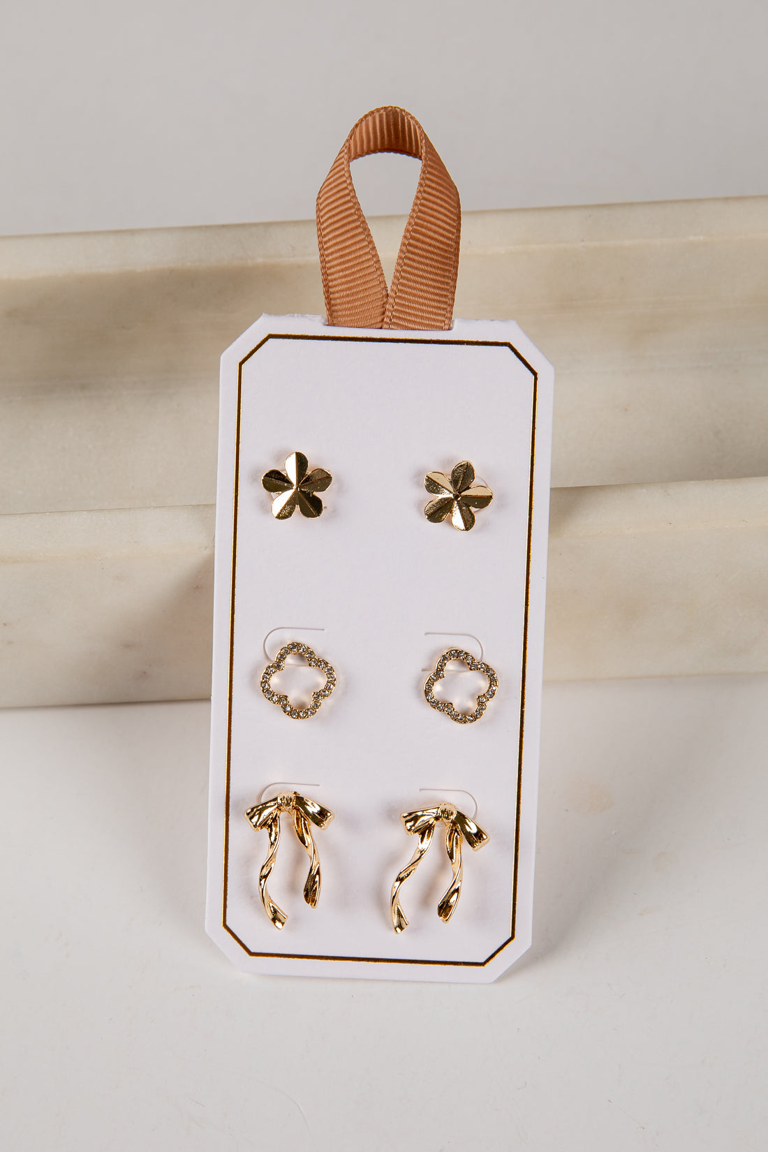 Set of gold earrings on a white card with a brown ribbon, against a neutral background.