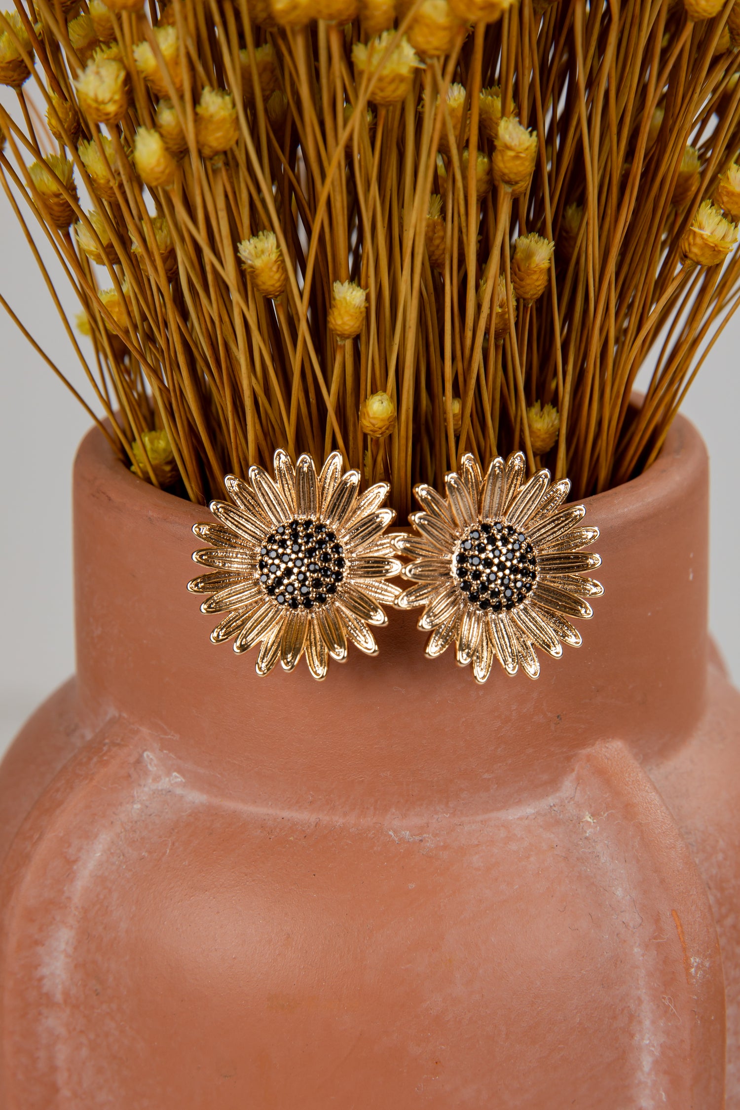 Gold sunflower earrings on a terracotta vase with dried flowers.