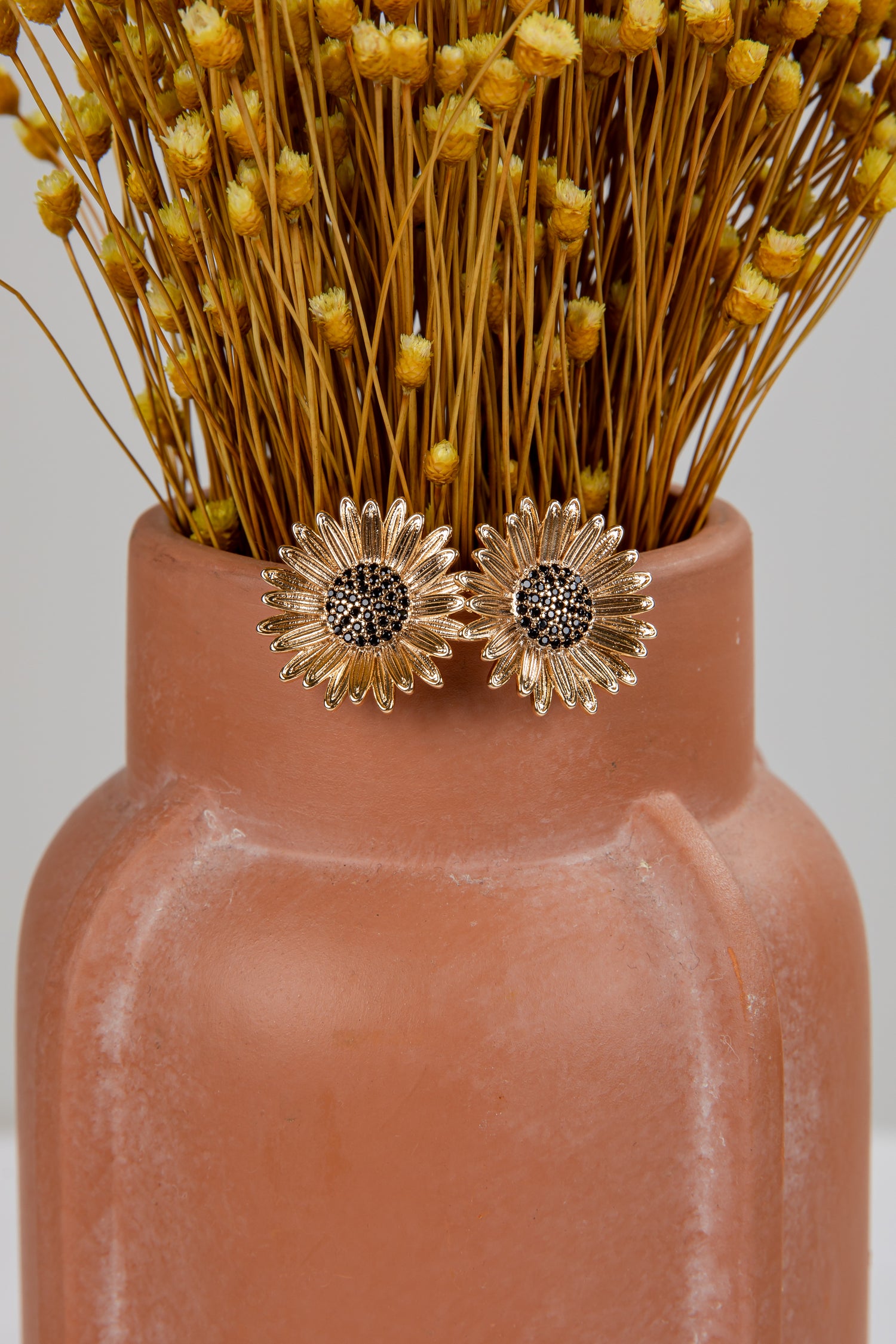 Gold sunflower earrings on a terracotta vase with dried flowers.