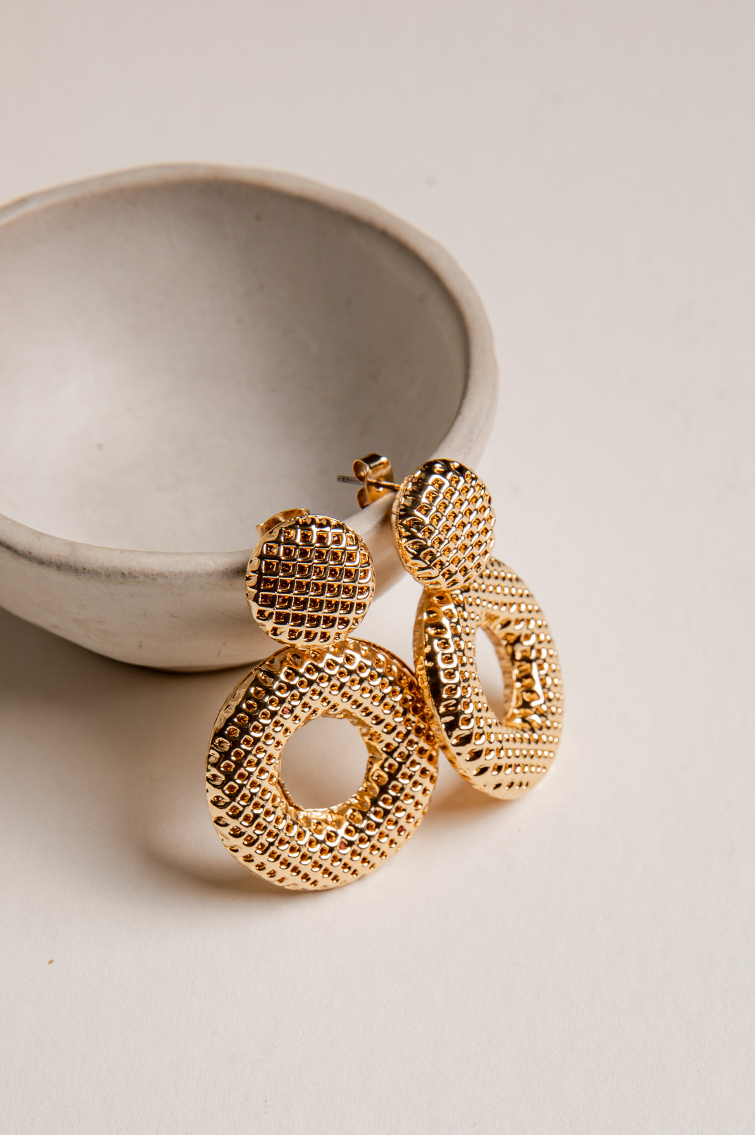 Gold hoop earrings on a light beige surface with a ceramic bowl in the background
