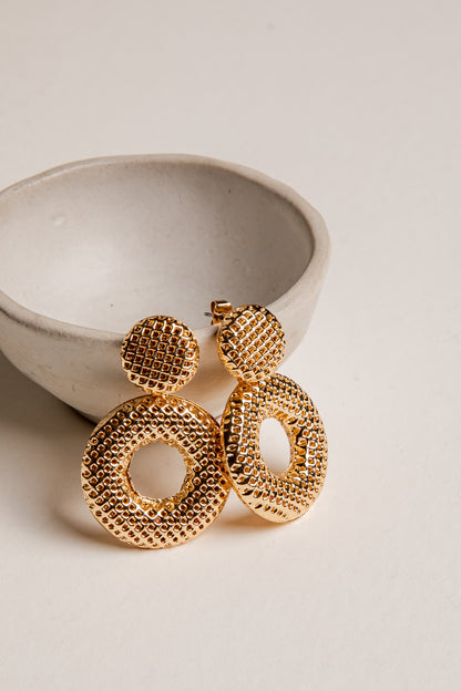 Gold earrings on a light beige surface with a ceramic bowl in the background