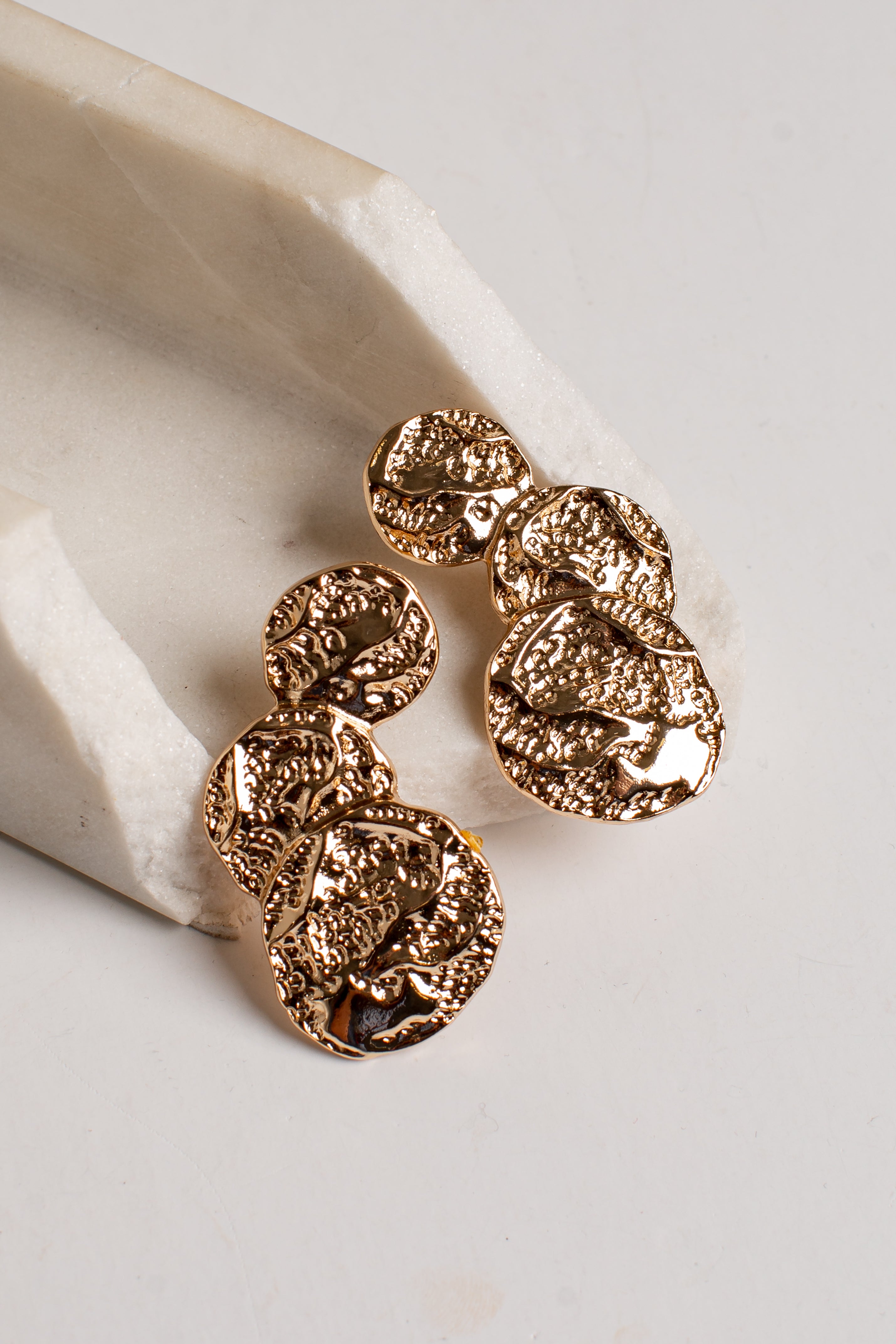 A pair of textured gold drop earrings with organic, coin-like shapes, displayed on a light beige stone prop against a neutral off-white background.
