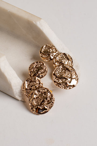 A pair of textured gold drop earrings with organic, coin-like shapes, displayed on a light beige stone prop against a neutral off-white background.