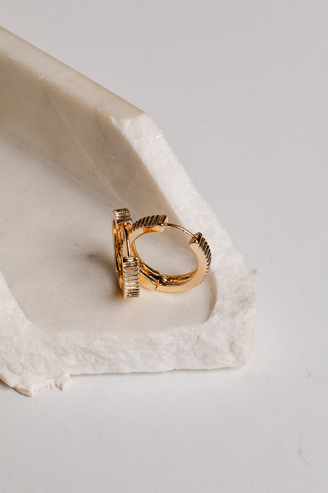 A gold huggie hoop ring with a textured twisted band and a small rectangular gold accent on the side, displayed on a white textured stone tray against a light gray background.