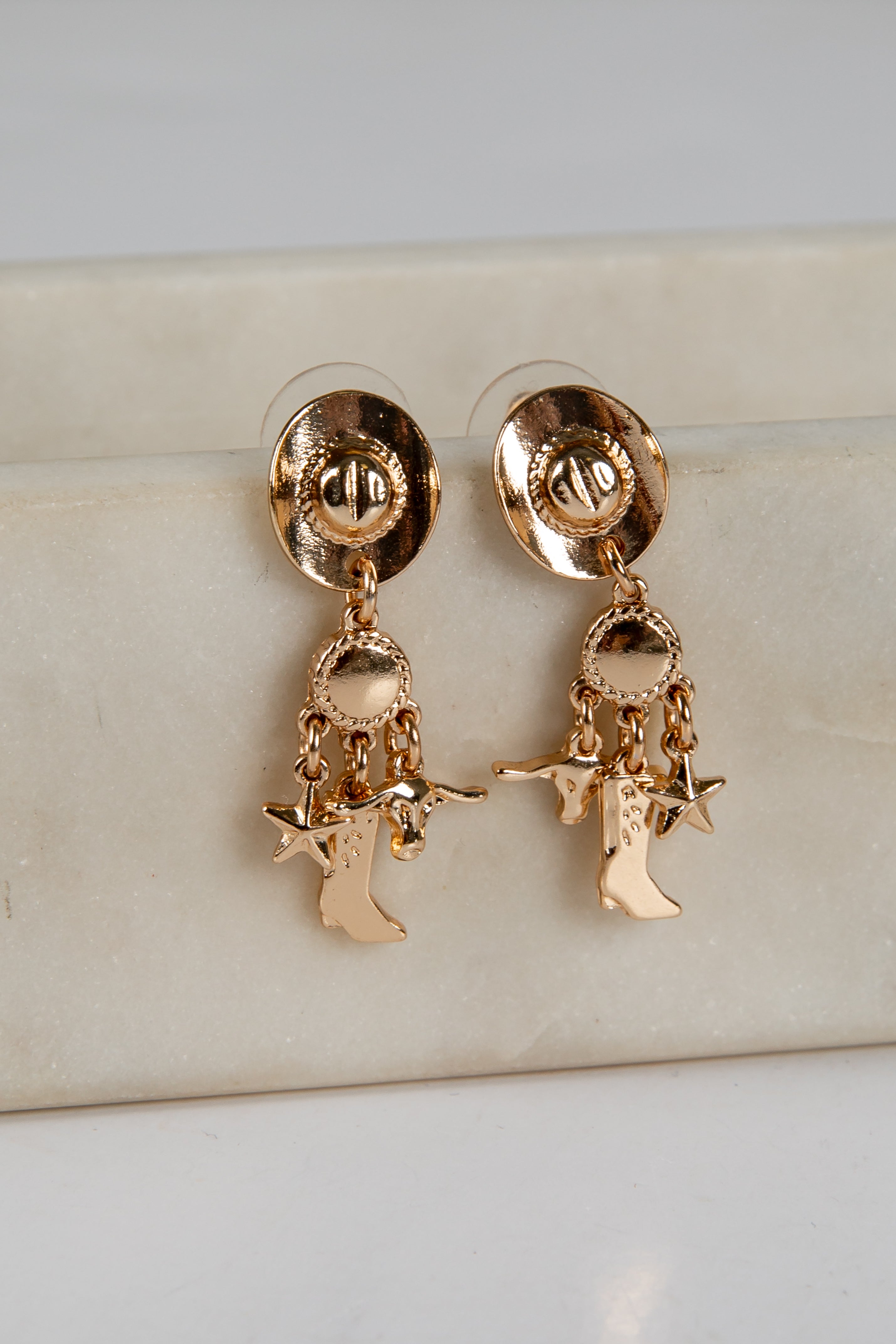 Gold earrings with cowboy boot and star charms on a beige surface.